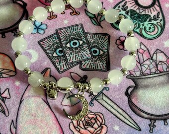 May include: A white beaded bracelet with silver accents and a crescent moon charm. The bracelet features round, opaque white beads and small silver beads. The charm is encrusted with small, clear crystals. The bracelet is set against a pink background with occult-themed illustrations.