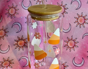 May include: A clear glass water bottle with a bamboo lid and a straw. The bottle is decorated with candy corn and flowers. The bottle is on a pink background with a sun and moon pattern.