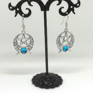 May include: Silver earrings with a pentacle design and a blue gemstone. The earrings are hanging from a black earring stand.