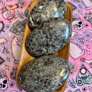 May include: Three smooth, oval-shaped stones with a speckled black and gray pattern. The stones are arranged in a wooden tray.