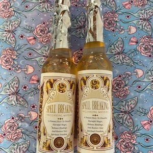 May include: Two glass bottles of "Spell Breaking Cleansing Water" with gold and cream labels. The bottles are sealed with silver foil and contain a light amber liquid. The background is a blue fabric with a floral and butterfly pattern.