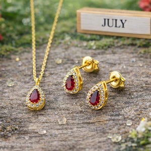 Puede incluir: Un conjunto de collar y pendientes dorados con piedras preciosas rojas en forma de lágrima y pequeñas piedras blancas. La palabra "JULY" se muestra en un bloque de madera. Las joyas están sobre una superficie de madera con flores rojas.