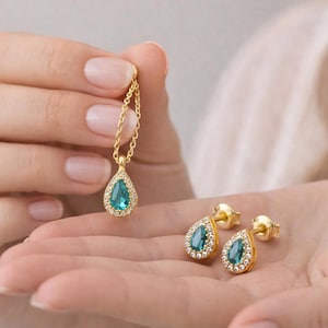 May include: A gold necklace and matching earrings set. The pendant and earrings feature a teardrop-shaped, teal-coloured stone surrounded by small, clear stones. The jewellery is held in a person's hand.