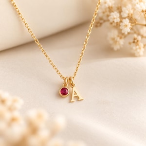 May include: A gold chain necklace featuring two small charms hanging from the center: a circular pendant set with a single round red gemstone and a capital letter A pendant. The necklace has a delicate link chain design with a polished finish.