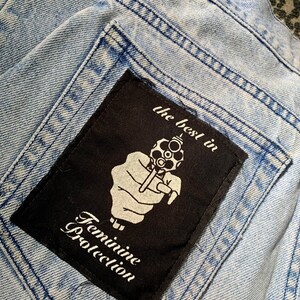 May include: A black patch sewn onto a light blue denim background. The patch features a white hand holding a gun with the text "the best in Feminine Protection" printed below.