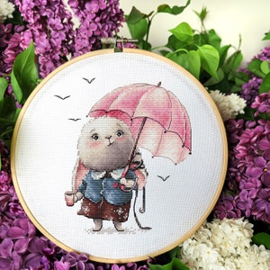 May include: A cross-stitch embroidery hoop with a cute cartoon bunny holding a pink umbrella and a cup. The bunny is wearing a blue jacket and a brown skirt. The background is white with a few birds flying.