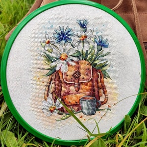 May include: A cross-stitch embroidery in a green hoop features a brown backpack with white daisies and blue flowers. A small gray mug is next to the backpack. A brown backpack is in the background.