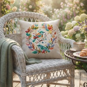 May include: A square throw pillow with a beige fabric cover featuring a colorful printed wreath of leaves and two small birds. The pillow rests on a white wicker chair draped with a sage green throw blanket, next to a small table with a croissant.