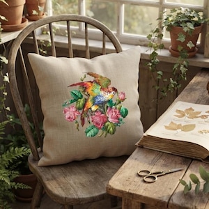 May include: A square beige throw pillow featuring a central graphic print of a colorful parrot perched among pink roses and green leaves. The pillow is made of a textured fabric and is positioned on a wooden chair in a sunlit room.