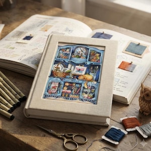 May include: A beige fabric-covered book featuring a central illustration of a blue hutch filled with craft supplies, a cat, and a butterfly painting. The book rests on a wooden surface alongside black pens, metal scissors, thread spools, and an owl.