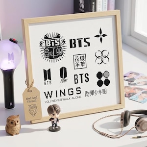 May include: A square framed cross-stitch pattern featuring black BTS-themed logos, symbols, and text including WINGS and YOU NEVER WALK ALONE. The design is displayed in a light wood frame with a tan tag labeled Owl And Thread attached to the side.