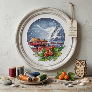 May include: A circular cross-stitch embroidery in a white wooden frame depicting a red house by a snowy mountain lake with orange cloudberries. A tag reads OWL AND THREAD COLLECTION NORDIC RETREAT SCANDI DREAMS SERIES. Spools of thread and scissors.