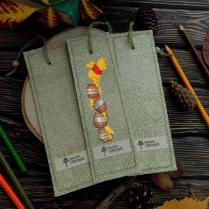 May include: Three green bookmark with a Winnie the Pooh cross-stitch design. The bookmark features Winnie the Pooh standing on top of a stack of honey pots. The bookmark has a label that says "Handmade".