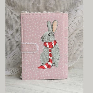 May include: A pink fabric notebook cover with a gray rabbit wearing a red and white scarf. The cover has a floral pattern.