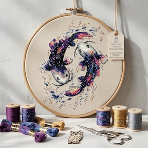 Pode incluir: Um bastidor de ponto cruz com dois peixes koi com padrões galácticos em roxo, azul e rosa. Uma etiqueta anexada diz OWL AND THREAD COLLECTION GALAXY KOI DANCE COSMIC JOURNEY SERIES. Inclui carretéis de linha e uma tesoura.