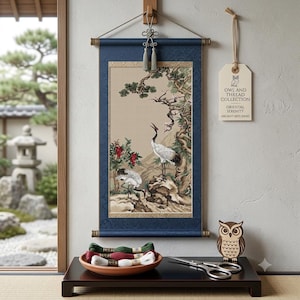 May include: A wall hanging scroll featuring a painting of two cranes, a pine tree, and red berries. A tag reads OWL AND THREAD COLLECTION ORIENTAL SERENITY ANCIENT ARTS SERIES. A wooden owl, scissors, and three skeins of embroidery thread are on a table.