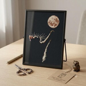 May include: A framed cross-stitch embroidery on black fabric featuring a textured, light-coloured moon and a stylised, abstract silhouette of a cat reaching upward. The piece is displayed in a thin black frame with a tag labelled Owl And Thread Art Edition.