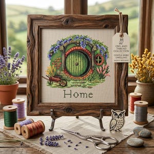 May include: A cross-stitch embroidery of a round green door dwelling with purple flowers and a wheelbarrow, framed in rustic wood. The text reads "Home" and "OWL AND THREAD COLLECTION GENTLE SHIRE DWELLING ANCIENT ARTS SERIES" on a hanging tag.
