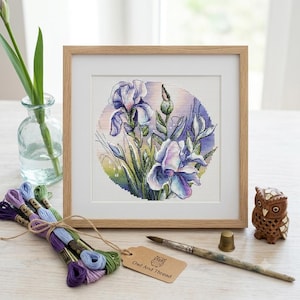 May include: A framed cross-stitch embroidery featuring purple and blue iris flowers on a circular background. The set includes a bundle of colourful embroidery floss, a wooden thimble, a paintbrush, and a tag labelled Owl And Thread.