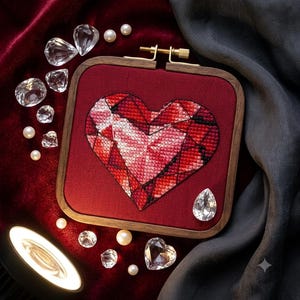 Può includere: Un telaio da ricamo quadrato in legno contenente un disegno a punto croce di un cuore sfaccettato nei toni del rosso, rosa e bianco. Il cuore è ricamato su uno sfondo di tessuto rosso scuro, incorniciato da un bordo in legno scuro con vite.