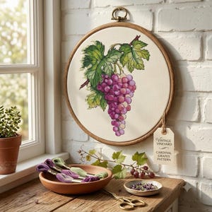 May include: A circular wooden embroidery hoop containing a cross-stitch design of a cluster of purple grapes with green leaves on cream fabric. A tag attached to the hoop reads Vintage Vineyard Cardinal Grapes Pattern, with embroidery floss and scissors.