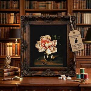 May include: A framed painting of a pale pink and white peony in a glass vase on a dark surface. A tan tag attached to the frame features an owl icon and the text: OWL AND THREAD COLLECTION, DUTCH MASTER PEONY PATTERN, WOODLAND TALES SERIES.