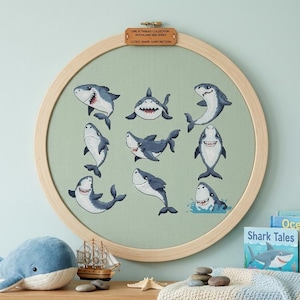 May include: A circular wooden embroidery hoop containing light green fabric with nine embroidered cartoon sharks in various poses. A small leather tag at the top reads "OWL & THREAD COLLECTION WOODLAND SEA SERIES LATEST SHARK GANG PATTERN".