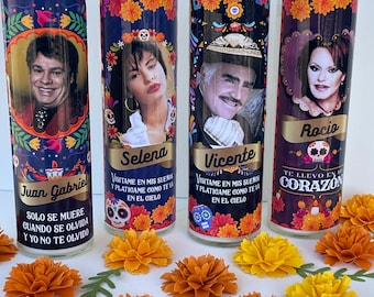 May include: Four glass prayer candles with portraits of Juan Gabriel, Selena, Vicente, and Rocio. Each candle has a colorful floral design and text in Spanish. Orange and yellow paper flowers are scattered in front.