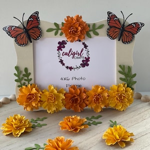 May include: A rectangular 4x6 photo frame with a light-colored border, decorated with two orange and black monarch butterfly cutouts, green leaf accents, and a row of layered paper marigold flowers in shades of yellow and orange.