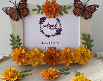 May include: A rectangular 4x6 photo frame with a light-colored border, decorated with two orange and black monarch butterfly cutouts, green leaf accents, and a row of layered paper marigold flowers in shades of yellow and orange.