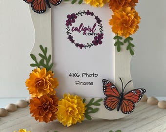 May include: A white 4x6 photo frame decorated with orange and yellow paper flowers, green paper leaves, and two monarch butterfly cutouts. The center of the frame features the text "caligirl CRAFTS" and "4x6 Photo Frame" printed on a white background.