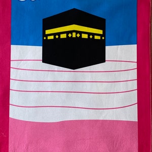 May include: A pink and blue prayer rug with a white background. The rug features a black and yellow image of the Kaaba, a white cloud, and a yellow sun. The rug has a pink border and silver fringe.