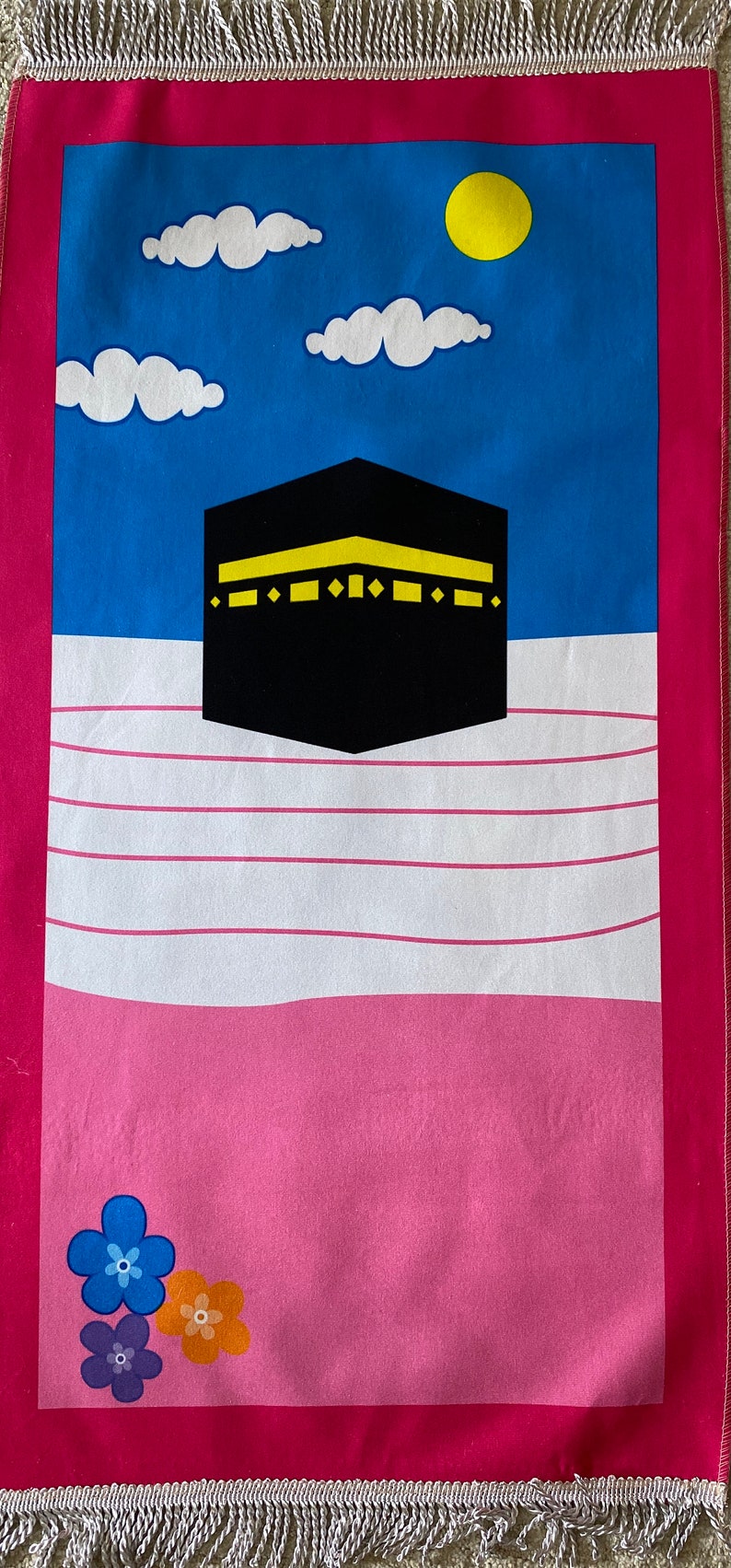 May include: A pink and blue prayer rug with a white background. The rug features a black and yellow image of the Kaaba, a white cloud, and a yellow sun. The rug has a pink border and silver fringe.