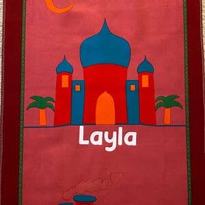 May include: A red, pink, and blue children's prayer rug with a mosque, clouds, a crescent moon, and a train. The rug has the name "Layla" written on it.