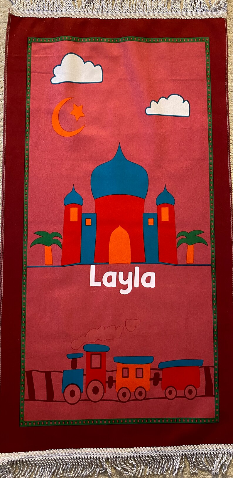 May include: A red, pink, and blue children's prayer rug with a mosque, clouds, a crescent moon, and a train. The rug has the name "Layla" written on it.