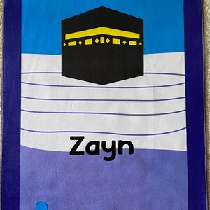 May include: A blue and purple prayer rug with a black and yellow image of the Kaaba, a white cloud design, and the name "Zayn" in black lettering. The rug has a fringe border.