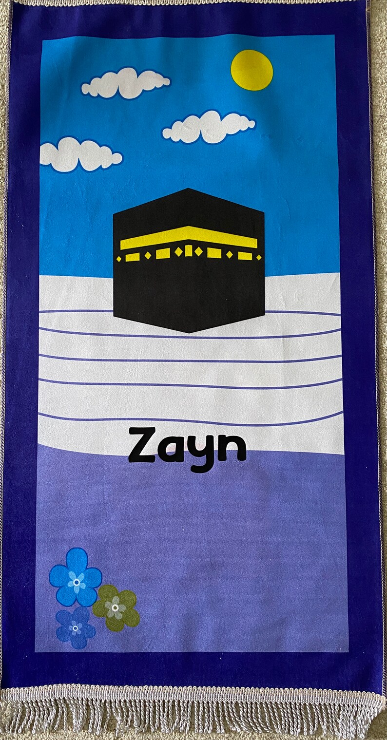 May include: A blue and purple prayer rug with a black and yellow image of the Kaaba, a white cloud design, and the name "Zayn" in black lettering. The rug has a fringe border.