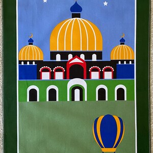 May include: A green prayer rug with a blue sky and white stars. A mosque with a yellow dome and blue minarets is in the center of the rug. A hot air balloon is flying above a green field with trees.