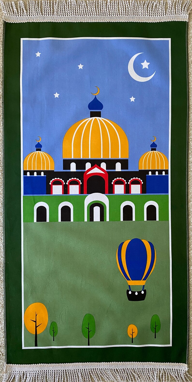May include: A green prayer rug with a blue sky and white stars. A mosque with a yellow dome and blue minarets is in the center of the rug. A hot air balloon is flying above a green field with trees.