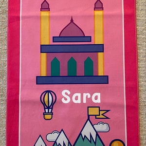 May include: Pink prayer rug with a mosque, stars, mountains, a hot air balloon, and the name "Sara" printed on it.