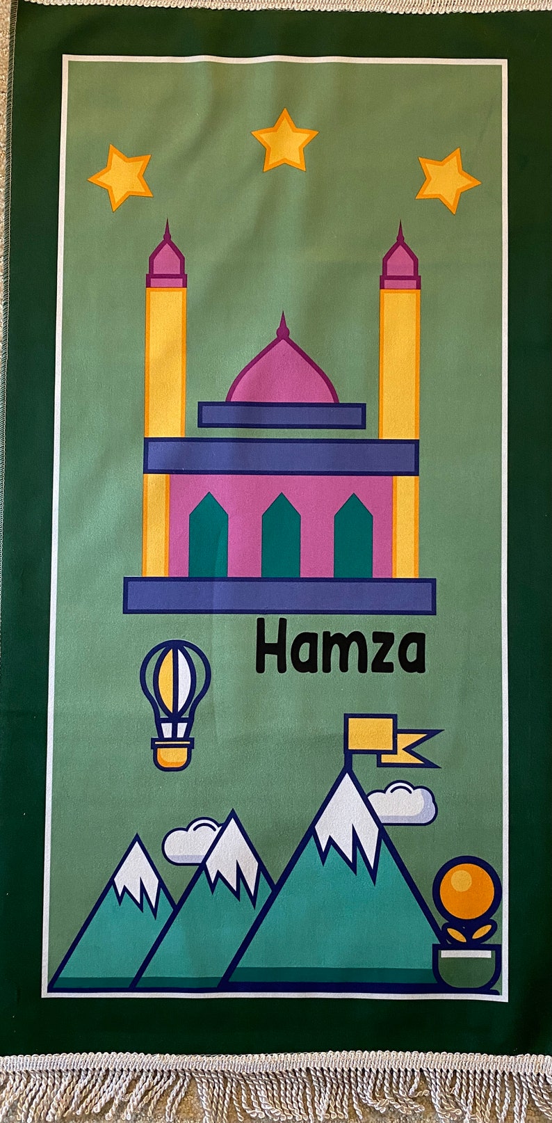 May include: A green prayer rug with a mosque, stars, mountains, a hot air balloon, and the name "Hamza" printed on it.