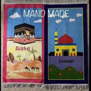 May include: Two handmade prayer rugs with colorful designs. The left rug features a pink background with a depiction of the Kaaba, a desert landscape, and the name Aisha. The right rug has a blue background with a mosque, green grass, and the name Ismael.