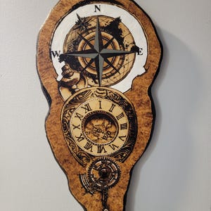 May include: Wooden wall art featuring a compass rose, a world map, and a clock design. The artwork is burned into a piece of wood with a natural edge. The compass rose has a black and gray design. The clock has Roman numerals.