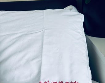 Pode incluir: Fronha branca com texto bordado a vermelho: "Night. Love you, sleep tight." A fronha está numa cama. Uma cabeceira escura e uma planta num vaso preto são visíveis no fundo.