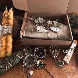 May include: A brown cardboard box filled with various items, including two yellow candles, a bundle of dried herbs, a tea strainer, a small wooden box, and two blue candles. The box is labelled "Mooncraft Magick".