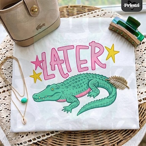 May include: White t-shirt featuring a cartoon alligator with the word "LATER" in pink bubble letters. The alligator is green with pink accents. Gold and turquoise jewelry, a purse, and a hair clip are also visible.