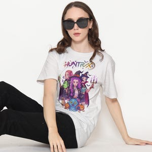 May include: White t-shirt with a colorful graphic featuring three cartoon witches, a blue cat, and pumpkins. The word "HUNTR" is printed above the graphic. The person in the image is wearing black sunglasses and black pants.