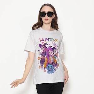 May include: White t-shirt featuring a colorful graphic of three stylized female figures in Halloween costumes, with the word "HUNTRX" above them. The design includes pumpkins, bats, and a blue cat.