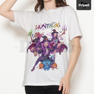 May include: White t-shirt featuring a colorful graphic of three witches, a blue cat, and pumpkins. The word "HUNTRX" is printed across the top. The witches are wearing hats and capes. The cat is wearing a witch hat. The shirt is made of a soft material.