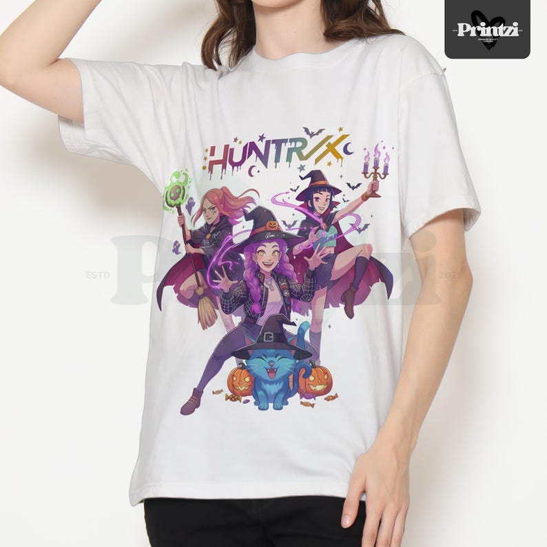 May include: White t-shirt featuring a colorful graphic of three witches, a blue cat, and pumpkins. The word "HUNTRX" is printed across the top. The witches are wearing hats and capes. The cat is wearing a witch hat. The shirt is made of a soft material.